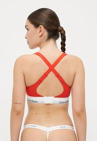 Červená sportovní podprsenka s křížovým designem, širokými ramínky a bílým elastickým páskem s logem "Calvin Klein". Hladká textilní struktura.