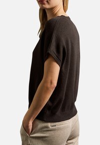 Donna che indossa un maglione a maniche corte, largo e scuro, e pantaloni beige chiaro, con le mani in tasca, mostrata di lato su uno sfondo bianco.
