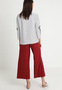 Gestreepte wit- en zwarte trui met driekwartmouwen en bloemengebreide; gecombineerd met ruime rode broeken. Eenvoudige platte schoenen zichtbaar.