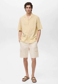 Korte mouwen, lichtgele blouse gemaakt van textuurstof, gecombineerd met beige cargoshorts en bruine sandalen.