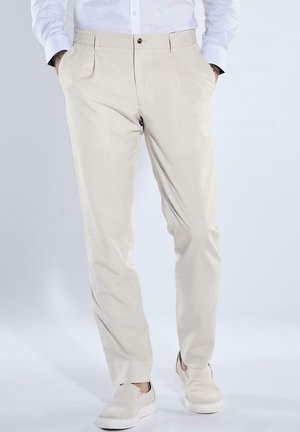 Man wearing beige slim-fit pants with side pockets and white slip-on shoes, standing with hands in pockets against plain background.