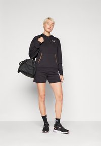 Sweat à capuche noir avec une poche avant, associé à un short noir. Accessoirisé avec un sac de sport noir doté de détails en maille et des chaussures de sport.