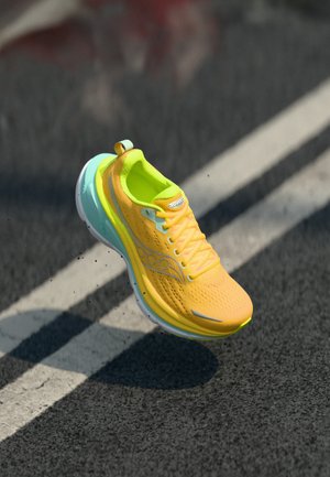 Chaussure de course jaune vif et bleu clair en l'air au-dessus d'une route en asphalte noire avec des marquages blancs.