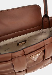 Borsa a mano in finta pelle marrone con design trapuntato, dotata di un interno beige chiaro con motivo ripetuto "GUESS" e una targhetta in metallo dorato.