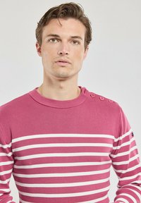 Pull rayé rose en matière côtelée, avec des rayures blanches horizontales, un col rond et trois boutons sur l'épaule gauche.