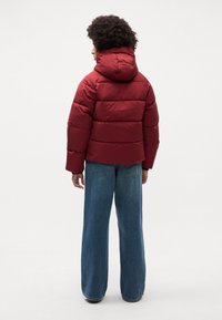 Chaqueta acolchada roja con capucha, diseño de rombos y puños elásticos, combinada con vaqueros de denim azul de corte holgado. Adecuada para el clima frío.