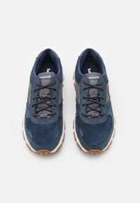 Mörkblå mockaskor med grå detaljer, texturerade snören och en vit mellansula. Har en gummisula med bruna mönster för grepp.