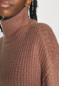 Pull à col roulé marron texturé avec un motif en maille épaisse. Il présente un col haut et une apparence douce et confortable avec une coupe décontractée.