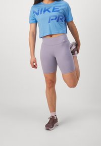 Blå kortärmad t-shirt med "NIKE PRO"-logotyp, matchad med ljuslila shorts. Modellen sträcker ut benet, iklädd bruna Nike-sneakers med rosa detaljer.