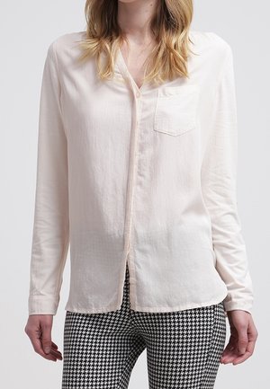 Blusa a maniche lunghe rosa chiaro con colletto, chiusura frontale a bottoni e taschino sul petto, abbinata a pantaloni a scacchi bianco e nero.