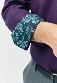 Fioletowa koszula z wzorzystym tealowym mankietem z motywem kwiatowym. Mankiet ma ostry kształt i jest wykonany z gładkiego materiału.