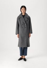 Cappotto grigio in lana con design doppiopetto, grandi revers, tasche frontali e bottoni, abbinato a jeans a gamba larga e scarpe slip-on nere.