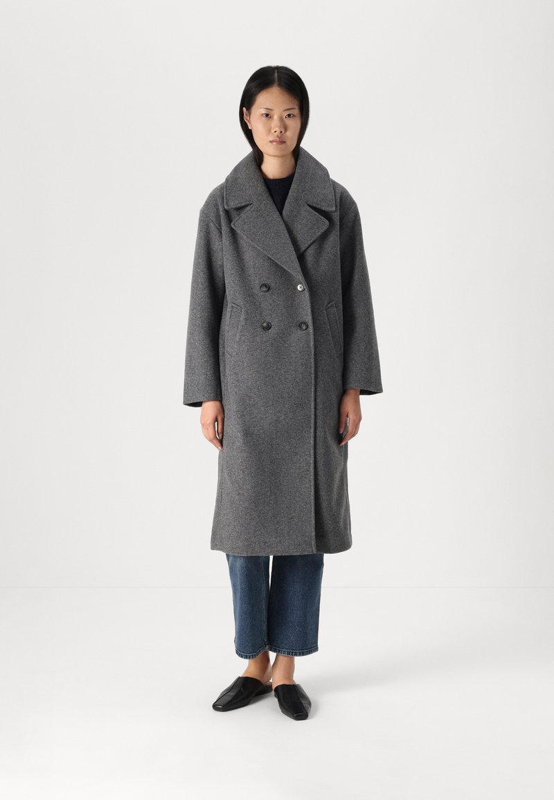 Cappotto grigio in lana con design doppiopetto, grandi revers, tasche frontali e bottoni, abbinato a jeans a gamba larga e scarpe slip-on nere.