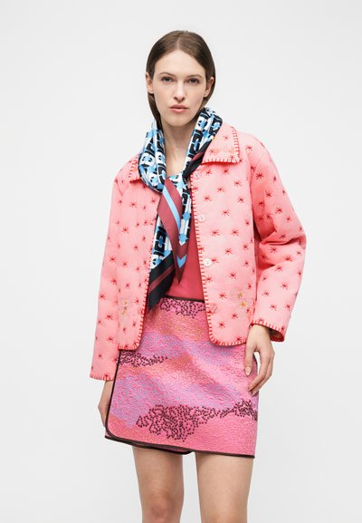 Γυναίκα που φοράει ροζ κεντημένο σακάκι, ροζ μίνι φούστα με σχέδια, μπλε και μαύρο φουλάρι με τύπωμα και κόκκινο τοπ μπροστά σε λευκό φόντο.