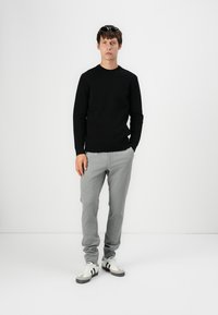 Pull en maille noir, pantalon à rayures grises, et sneakers blanches avec accents noirs. Le mannequin pose devant un fond blanc uni.