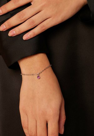 Silver chain bracelet with a round pink gemstone charm, worn on a wrist against a dark fabric background. Smooth texture and delicate design.