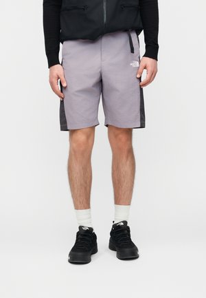 Άνδρας που φοράει ανοιχτό μωβ σορτς με μαύρες πλαϊνές λεπτομέρειες, μαύρα αθλητικά παπούτσια, λευκές κάλτσες και μαύρο σακάκι με τα χέρια στα πλευρά.