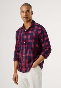 Camisa de botão xadrez em azul-marinho e vermelho, com colarinho espalhado, mangas compridas arroladas nos punhos e botões brancos. Tecido leve com uma textura suave.