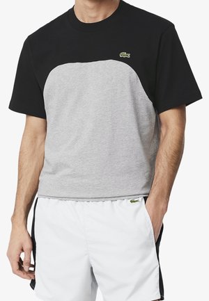 Homme portant un t-shirt à manches courtes noir et gris à blocs de couleur et un short blanc avec un petit logo crocodile vert sur les deux pièces.