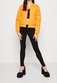 Chaqueta bomber acolchada naranja con tejido texturizado, cierre de cremallera y puños acanalados. Combinada con pantalones vaqueros skinny negros y tacones de plataforma negros.
