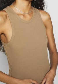 Top sin mangas de canalé marrón con amplias sisas y tejido texturizado. Presenta un escote redondo y una silueta ajustada. Collar plateado acentuado.