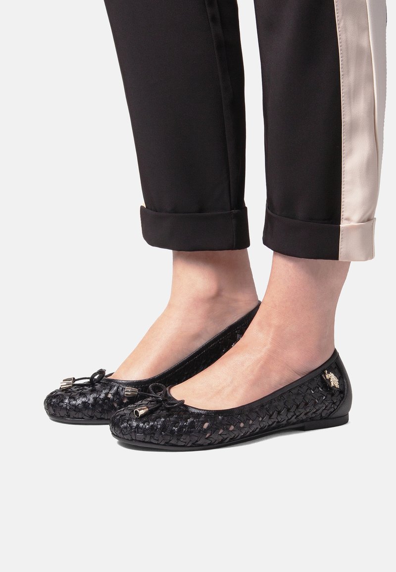 Fødder iklædt sorte vævede balletsko med små sløjfer, kombineret med sorte cropped bukser med en beige sideliste.