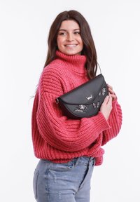 Černá dámská kabelka s výšivkou motýla a magnetickým uzávěrem. Měkká kůžová textura, polokruhový tvar, nošená v ruce vedle růžového svetru.