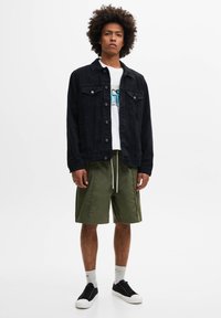 Jeune homme avec coiffure afro portant une veste en jean noire, un t-shirt blanc à motif, un short vert olive, des chaussettes blanches et des baskets noires, debout.