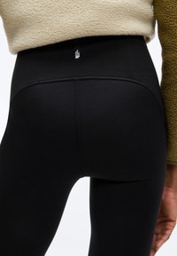 Κοντινό πλάνο ατόμου που φοράει μαύρα κολάν με ψηλή μέση και το λογότυπο της The North Face στην ζώνη μέσης, καθώς και μπλούζα από φλις σε μπεζ και ελιάς χρώμα.