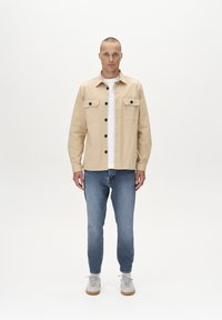 Beige gombos dzseki sötét gombokkal, két mellzsebbel, fehér póló fölött viselve, világoskék farmernadrággal és világos sportcipővel párosítva.