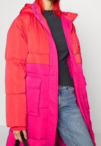 Manteau matelassé avec un design en blocs de couleur, comprenant des sections roses vives et corail, une capuche, une fermeture éclair au devant et une grande poche latérale.
