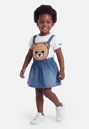Osmijehno dijete u bijeloj majici s velikom grafikom plišanog medvjedića i plavoj traper haljini s tregerima, bijele tenisice.