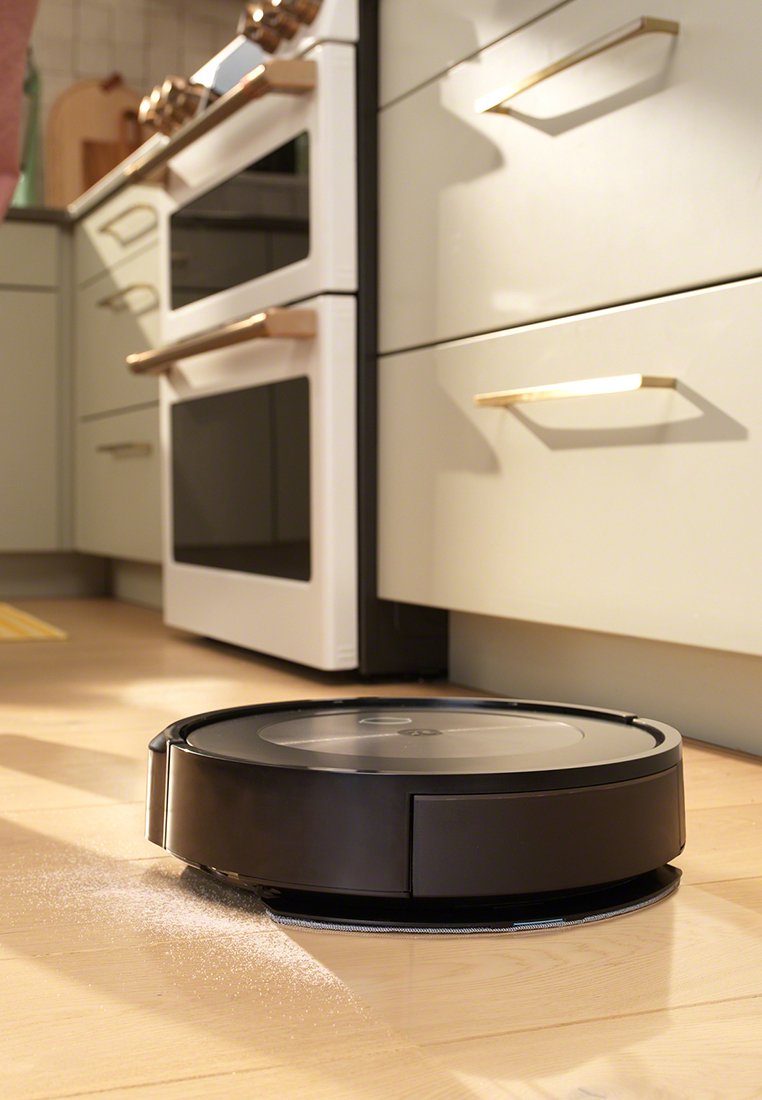 Ein schwarzer robotergestützter Staubsauger auf einem Holzfußboden in der Küche, in der Nähe eines Herdes und Schränken, mit sichtbarem Staubstreifen daneben.