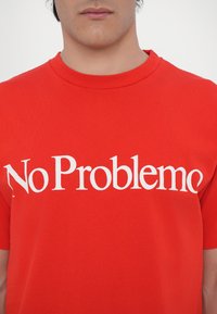 Rotes Baumwoll-T-Shirt mit kurzen Ärmeln und Rundhalsausschnitt, auf dem der Text "No Problemo" in großer, weißer Serifenschrift über dem Oberkörper abgedruckt ist.