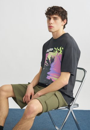 Jonge man met krullend haar, zittend op een metalen stoel, gekleed in een zwart T-shirt met afbeelding en olijfgroene cargobroek, tegen een effen achtergrond.