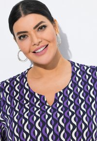 Top de color púrpura y blanco con un diseño de escote en V. Tela suave con patrones en ondas. La modelo lleva grandes pendientes de aro plateados.