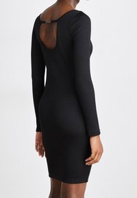 Robe noire côtelée avec manches longues et découpe au dos ornée de ferrures métalliques. Le tissu présente une texture lisse et une silhouette ajustée.