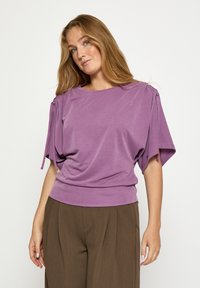 Lavender purple shirt with loose, short sleeves, gathered details at shoulders, and a fitted bottom hem, paired with brown wide-leg trousers.