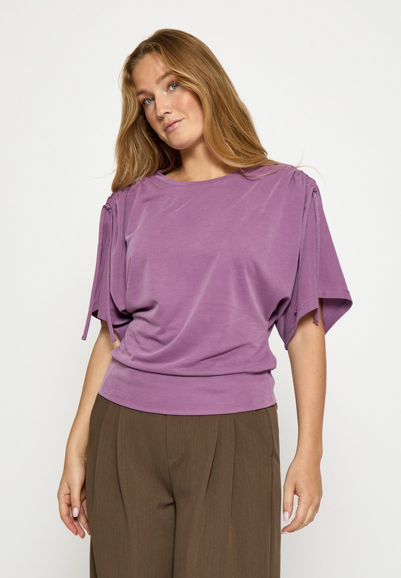 Lavender purple shirt with loose, short sleeves, gathered details at shoulders, and a fitted bottom hem, paired with brown wide-leg trousers.