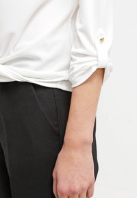 Camicia bianca a maniche lunghe con polsini arrotolati, caratterizzata da un piccolo bottone a forma di cuore, abbinata a pantaloni neri, che mostrano una trama di tessuto liscia.