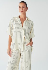 Vrouw met vastgebonden haar, gekleed in een roomkleurig los shirt en broek met zwarte geometrische lijnpatronen, met één hand in de zak.