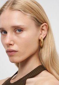 Blonde Frau mit blauen Augen, die große goldene Creolen und ein braunes gestricktes Oberteil trägt, schaut leicht zur Seite vor einem weißen Hintergrund.