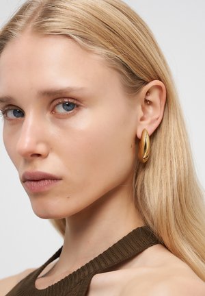 Blonde Frau mit blauen Augen, die große goldene Creolen und ein braunes gestricktes Oberteil trägt, schaut leicht zur Seite vor einem weißen Hintergrund.