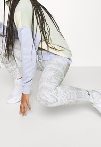 Person med långa flätor som bär en pastellfärgad färgblockertröja, marmorleggings och vita sneakers, sitter på huk med en hand på golvet.