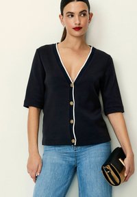 Marineblå cardigan med dyb V-hals, hvid kant og gyldne knapper. Korte ærmer og tætsiddende design, parret med lys blå jeans og en sort clutch.