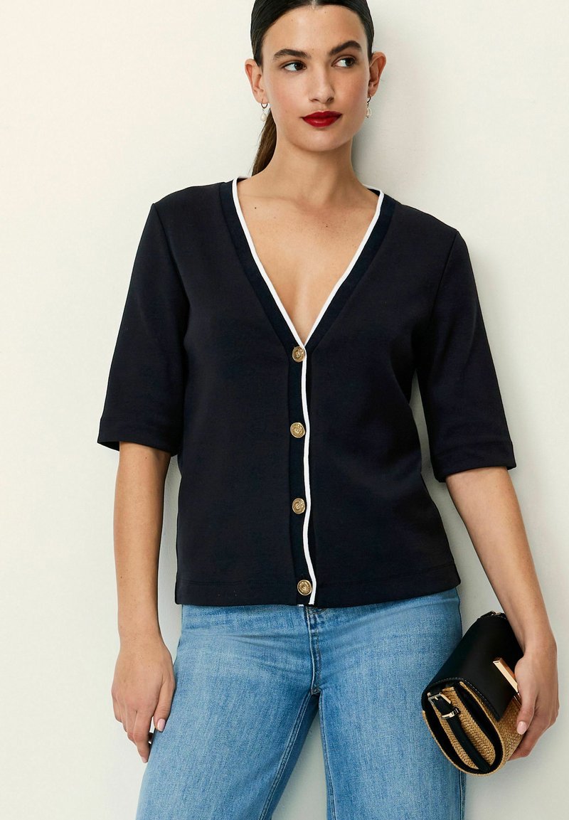 Marineblå cardigan med dyb V-hals, hvid kant og gyldne knapper. Korte ærmer og tætsiddende design, parret med lys blå jeans og en sort clutch.