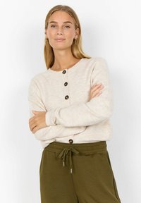 Blonde woman with long hair wears cream buttoned cardigan and olive green drawstring pants, standing with arms crossed against white background.