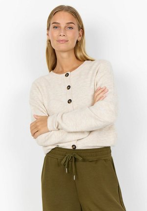 Blonde woman with long hair wears cream buttoned cardigan and olive green drawstring pants, standing with arms crossed against white background.