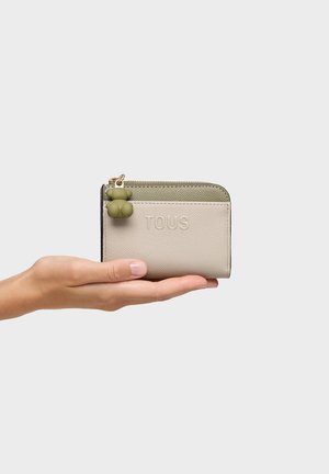 Ręka trzymająca mały beżowo-oliwkowy portfel TOUS z zamkiem błyskawicznym i zielonym suwakiem w kształcie misia.