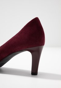 Burgundyröd mocka pumps med medelhög klack, slät och smal klack samt rundad bakdel på vit yta och ljusgrå bakgrund.